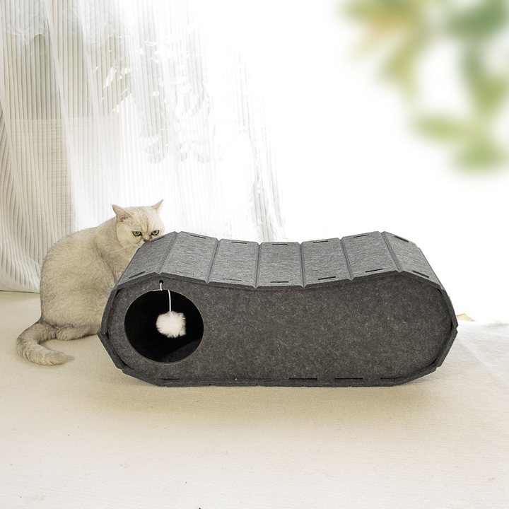 Dark grey felt sloped cat tunnel bed with a hanging pompom toy, shown in a room setting with a cat beside it.
