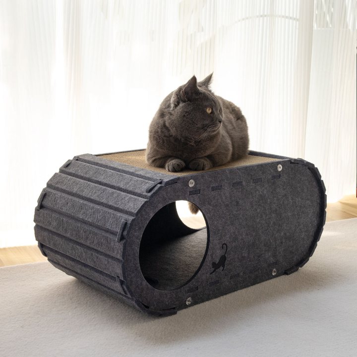 Dark grey felt capsule-shaped cat tunnel bed with a corrugated cardboard top pad, shown with a cat resting on top in a room setting.