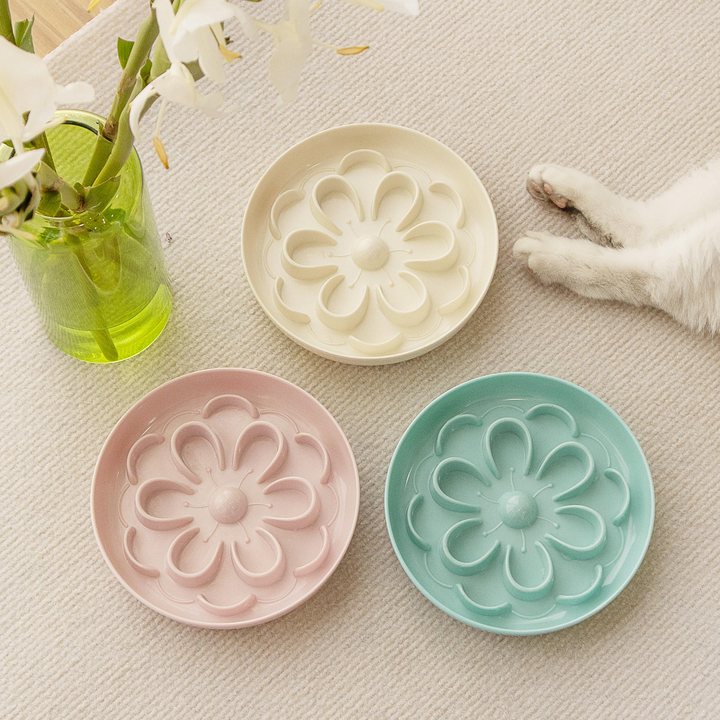 Flower-shaped pet slow feeder bowls in cream, pink, and teal, styled on a textured mat