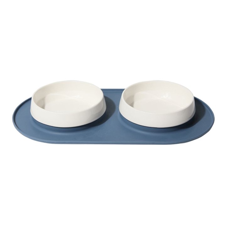 Two white ceramic pet bowls set into an oval blue-grey silicone mat