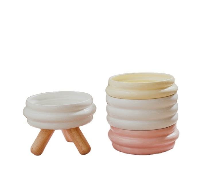 White ribbed ceramic pet bowl on wooden tripod legs, beside a stack of matching bowls in other colours