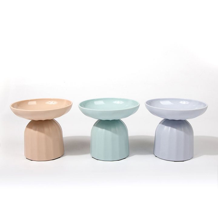 Three elevated ceramic pet bowls with a fluted pedestal base, in peach, mint green, and lavender.
