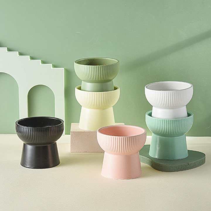 Elevated ceramic pet bowls with a fluted pedestal base, shown in black, pink, yellow, cream, white, and mint, styled against a green backdrop with a cut-out arch and wooden block.