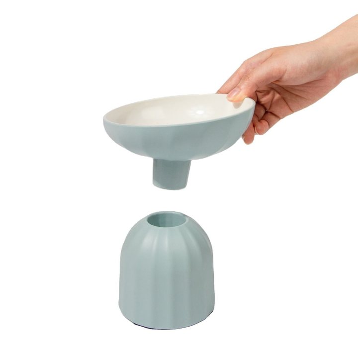 Two-piece round pet feeding bowl set in sage-green plastic with a white interior: a shallow tilted upper bowl on a short cylindrical stem being lifted by a hand, shown above its matching domed base with a circular opening, against a plain white background.