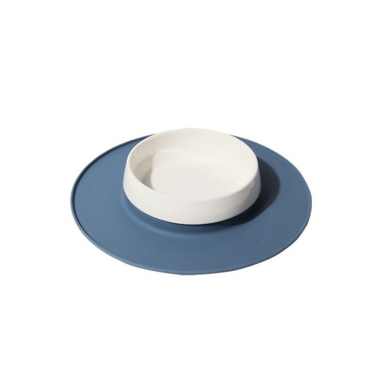 Round blue silicone pet placemat with a raised rim, topped by a white ceramic pet bowl with a curved interior divider.