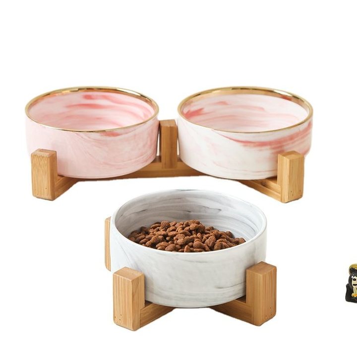 Two pink-and-white marble-effect ceramic bowls with gold rims set in a wooden frame stand, with a third matching bowl filled with kibble.
