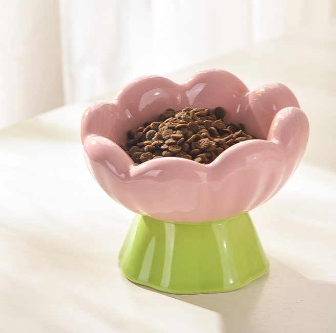 Pink flower-shaped ceramic pet bowl on a green pedestal foot, filled with kibble, on a sunlit tabletop.