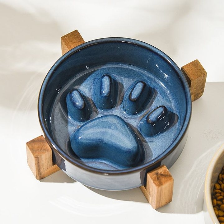 Blue ceramic slow feeder bowl with a moulded paw-print relief inside, set on a wooden four-leg stand on a sunlit surface.