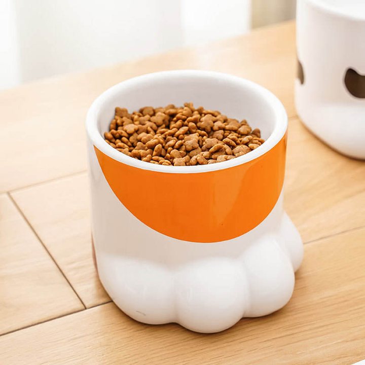 White and orange ceramic bowl moulded in the shape of a cat's paw, filled with kibble, on a wooden tabletop.