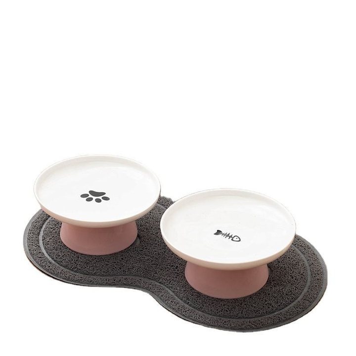Two round white ceramic pet bowls, each raised on a short pink cylindrical pedestal of slightly different heights, arranged side by side on a grey bone-shaped non-slip mat with a raised drainage-groove texture; one bowl's interior has a small paw-print design, the other a small fish-skeleton design.