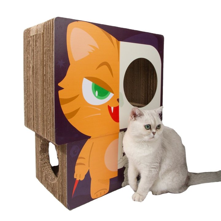 A square corrugated-cardboard cat house/scratcher with a large cartoon orange-cat face graphic on the front panel and a round entry hole on the top-front face, with a light grey shorthair cat sitting beside it.