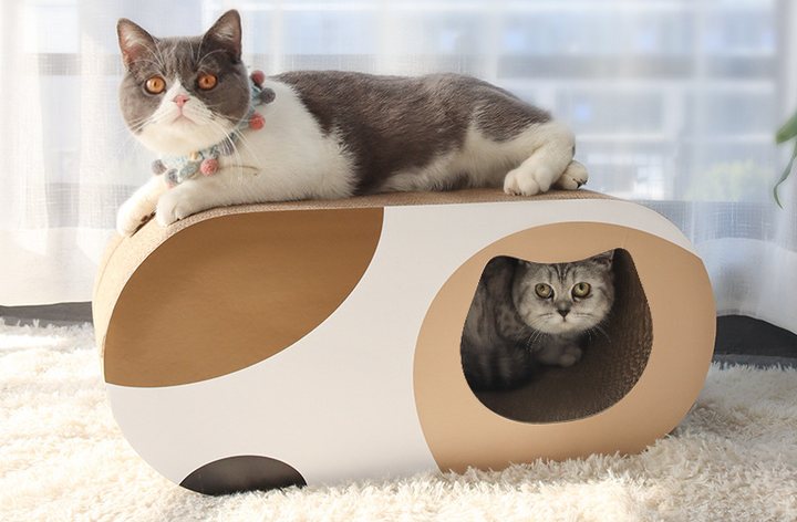 Oval cat scratcher lounge with a hollow tunnel through the body, in cream and tan corrugated board, one cat resting on the curved top and a second cat looking out of the tunnel opening.