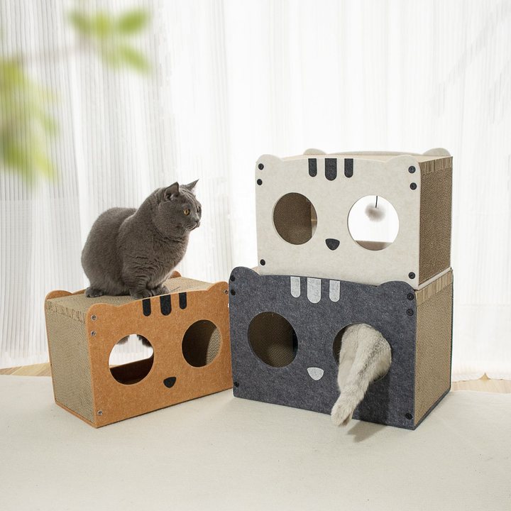 Cat-face shaped cardboard scratcher houses in tan, cream and dark grey felt, stacked in a living room with a hanging ball toy accessory