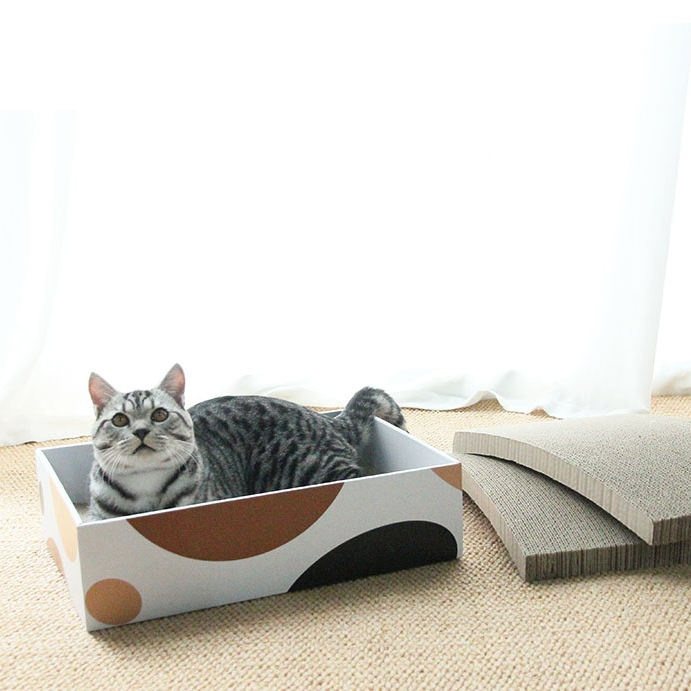 white/orange-graphic cardboard cat scratcher box with a grey tabby cat sitting inside it, two spare corrugated refill pads beside it
