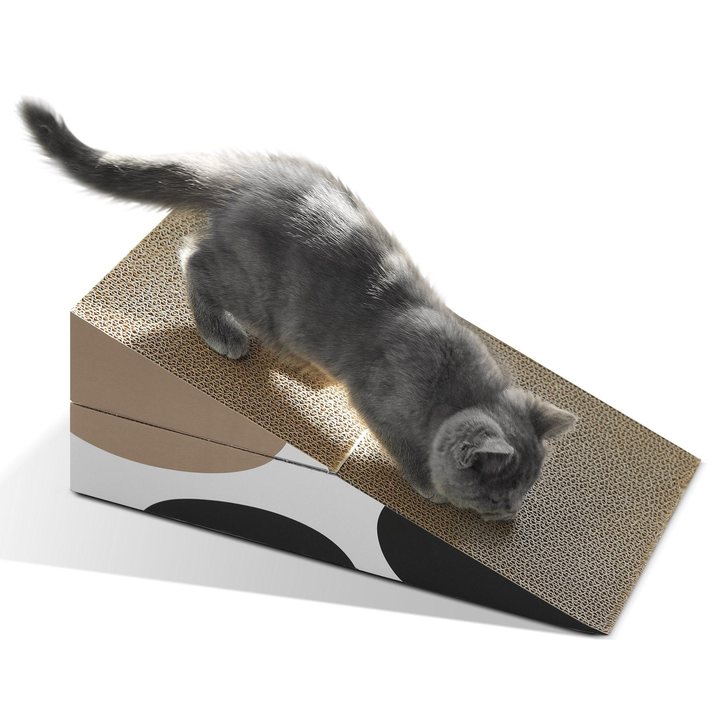 A grey long-haired kitten reaching down the sloped, corrugated-cardboard surface of a wedge-shaped cat scratcher; the scratcher has a tan corrugated top and a white side panel printed with a black and grey oval pattern.