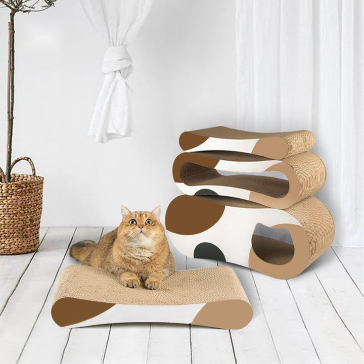 A tiered wave-shaped corrugated cardboard cat scratcher in cream and tan with brown accent patches, beside a flat rectangular cardboard scratch pad on the floor where a ginger cat is lying, set against a white wall with a sheer curtain and a potted tree in a woven basket.
