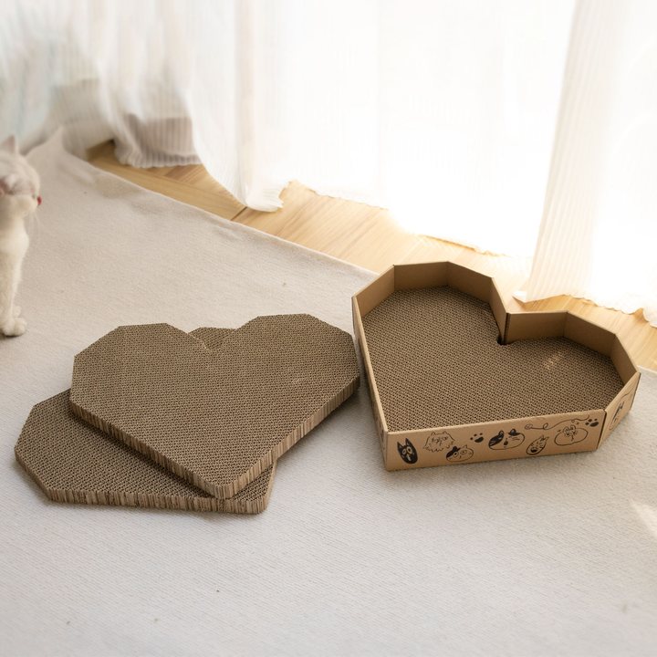 Heart-shaped corrugated cardboard cat scratch pads and a matching heart-shaped tray with a printed cat-doodle border, set on a floor rug.