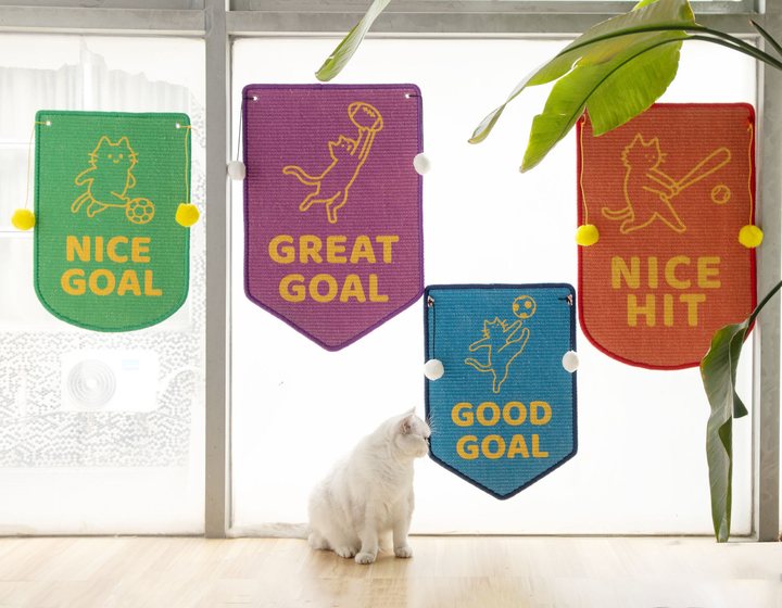 Flag-shaped felt cat scratch mats printed with sports-themed cat graphics, hung with pompom tassels on a window.