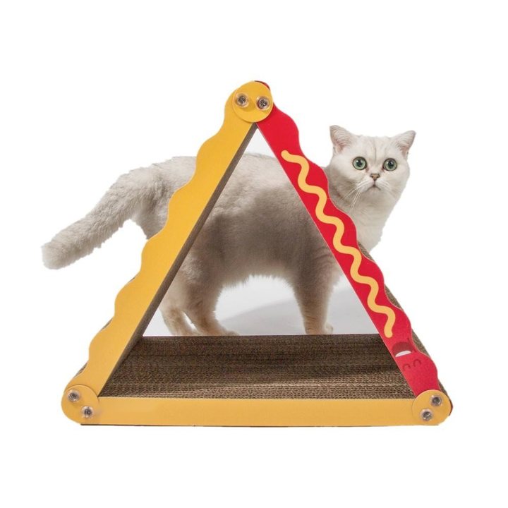 A triangular cardboard cat scratcher shaped like a hot dog, with a yellow corrugated-cardboard "bun" frame and a red panel with a mustard-yellow squiggle design, standing on a corrugated cardboard base; a light gray-and-white shorthair cat stands inside the triangular frame.