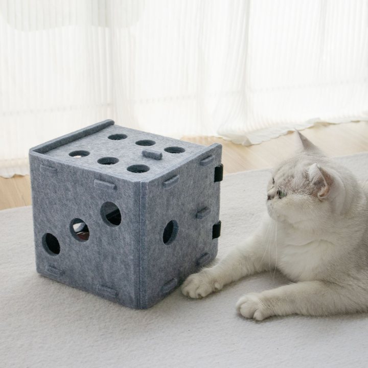 Grey felt dice-shaped cat puzzle box with circular cut-out holes, shown beside a resting cat.